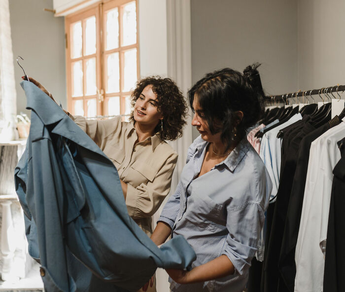 Two women in a store looking at a blue shirt on a hanger. This image relates to clothing sizes and dress experiments. Two women in a store looking at a blue shirt on a hanger. This image relates to clothing sizes and dress experiments.
