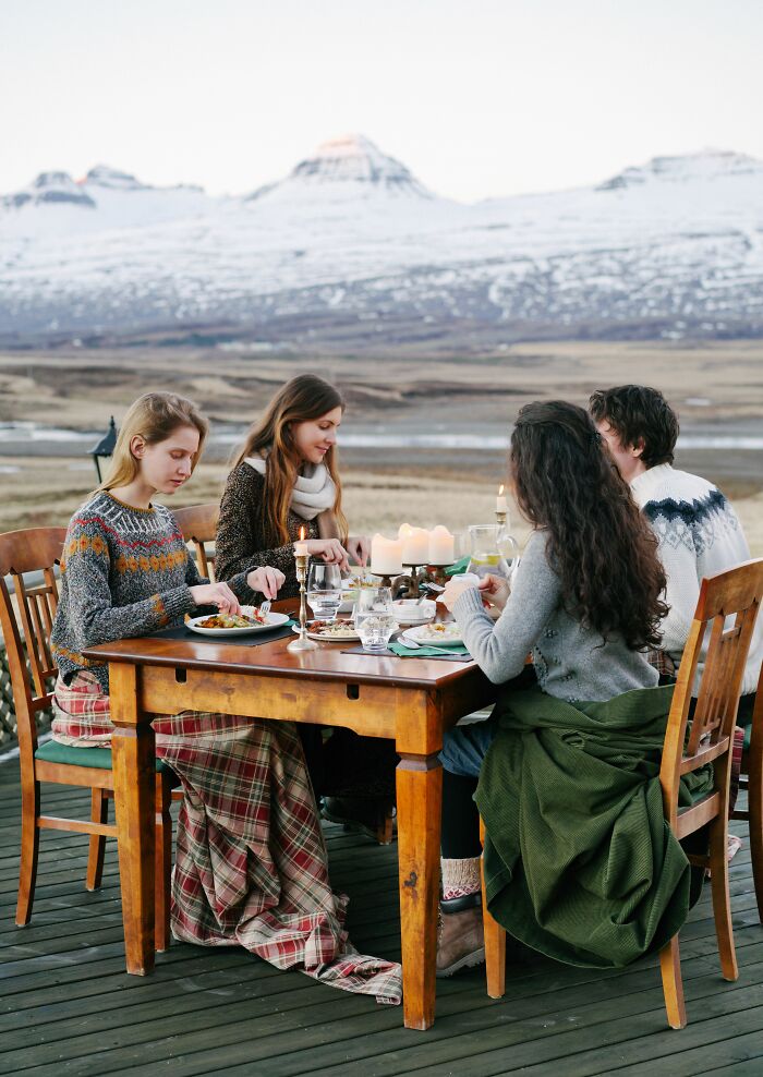 Four individuals, a mix of locals and foreigners, dine outdoors with snowy mountains in the background, a setting for cultural misunderstandings.