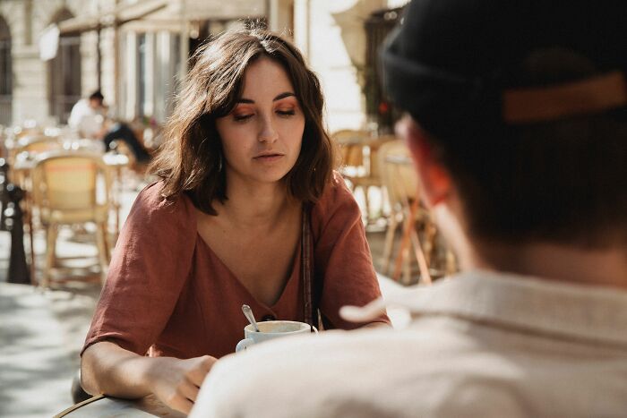 A woman with brown hair in a pink top looks down at a coffee cup in an outdoor cafe. This shows women changing their minds.