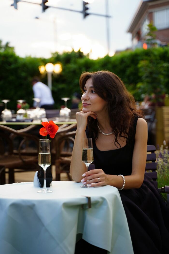 A elegant woman in a black dress and pearls, holding a champagne glass at an outdoor table, revealing true colors.