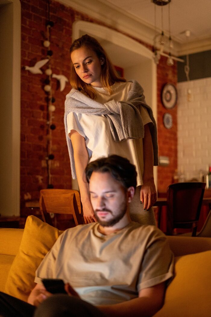 A woman stands behind a man engrossed in his phone, showing cheating red flags of emotional distance.
