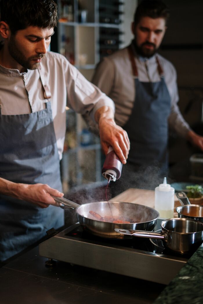 Two chefs in aprons cook in a kitchen, one adding sauce to a pan, illustrating a unique work story.