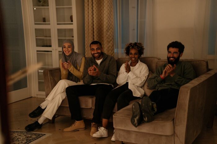 Four diverse friends happily clapping while watching TV together on a couch, showing genuine friendship.