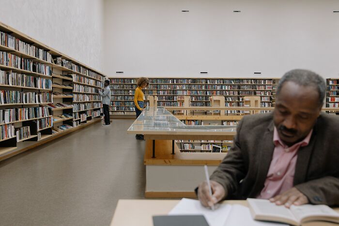 A man studies intently for a general knowledge quiz in a library, surrounded by bookshelves and other patrons.