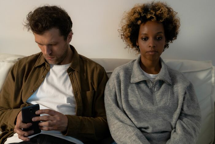 A couple sits apart on a couch, the man on his phone, highlighting cheating red flags of disconnection.