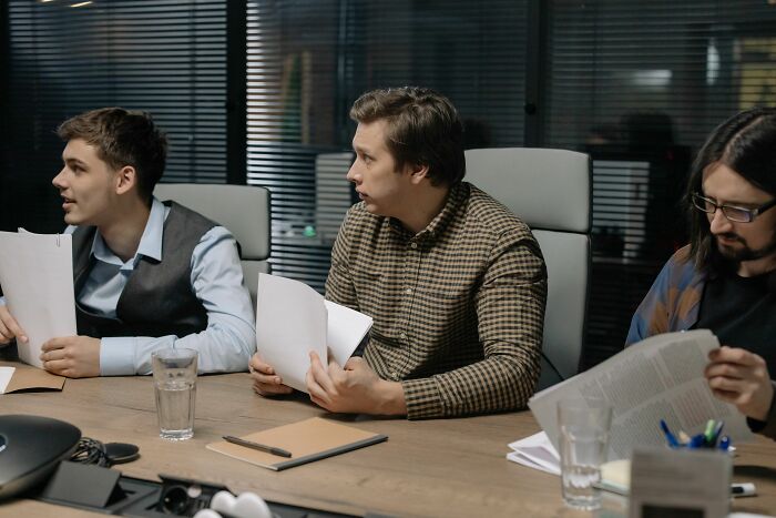 Three men are seated at a conference table, involved in an office story.