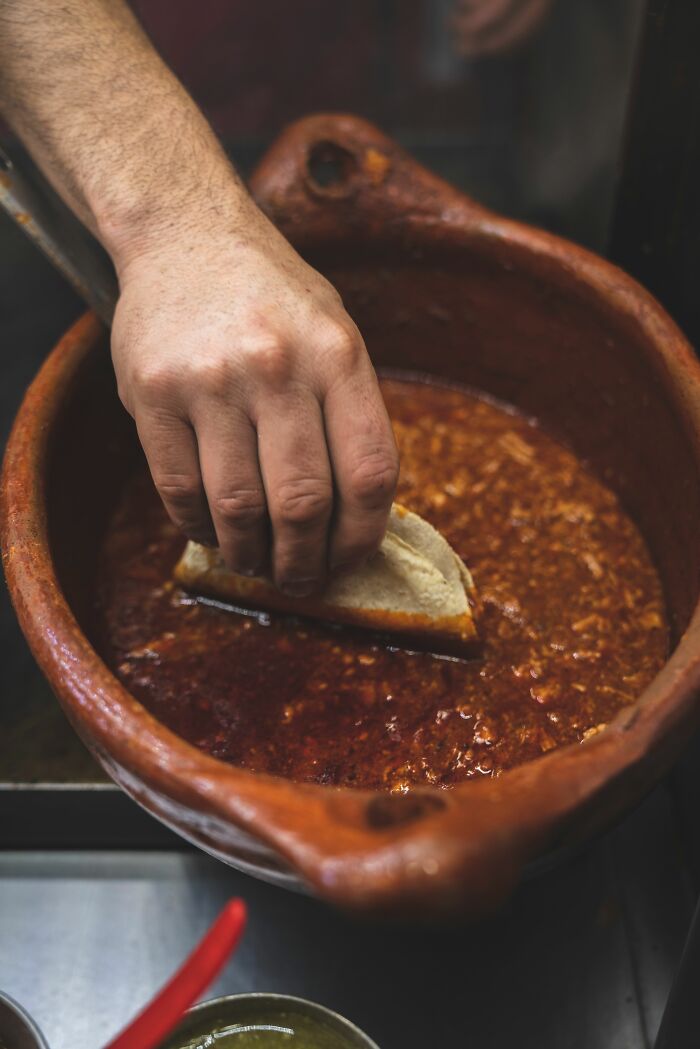 Hand dipping a taco into a large pot of stew, hinting at culinary practices that could fail health inspections.