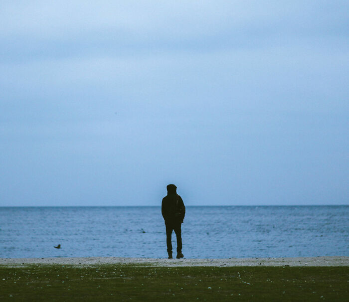 A lonely man stands by the vast ocean, facing the water under a somber sky, representing deep loneliness and isolation. A lonely man stands by the vast ocean, facing the water under a somber sky, representing deep loneliness and isolation.