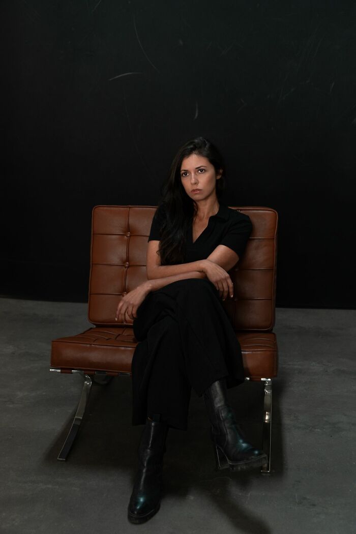 A woman in a black dress and boots sits on a brown leather chair against a dark background, reflecting the diverse origins of 'white phrases'.