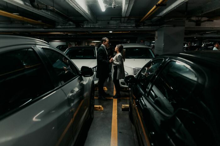 Two people talk between parked cars in a dimly lit underground parking garage. This image relates to secrets and how they could do damage.