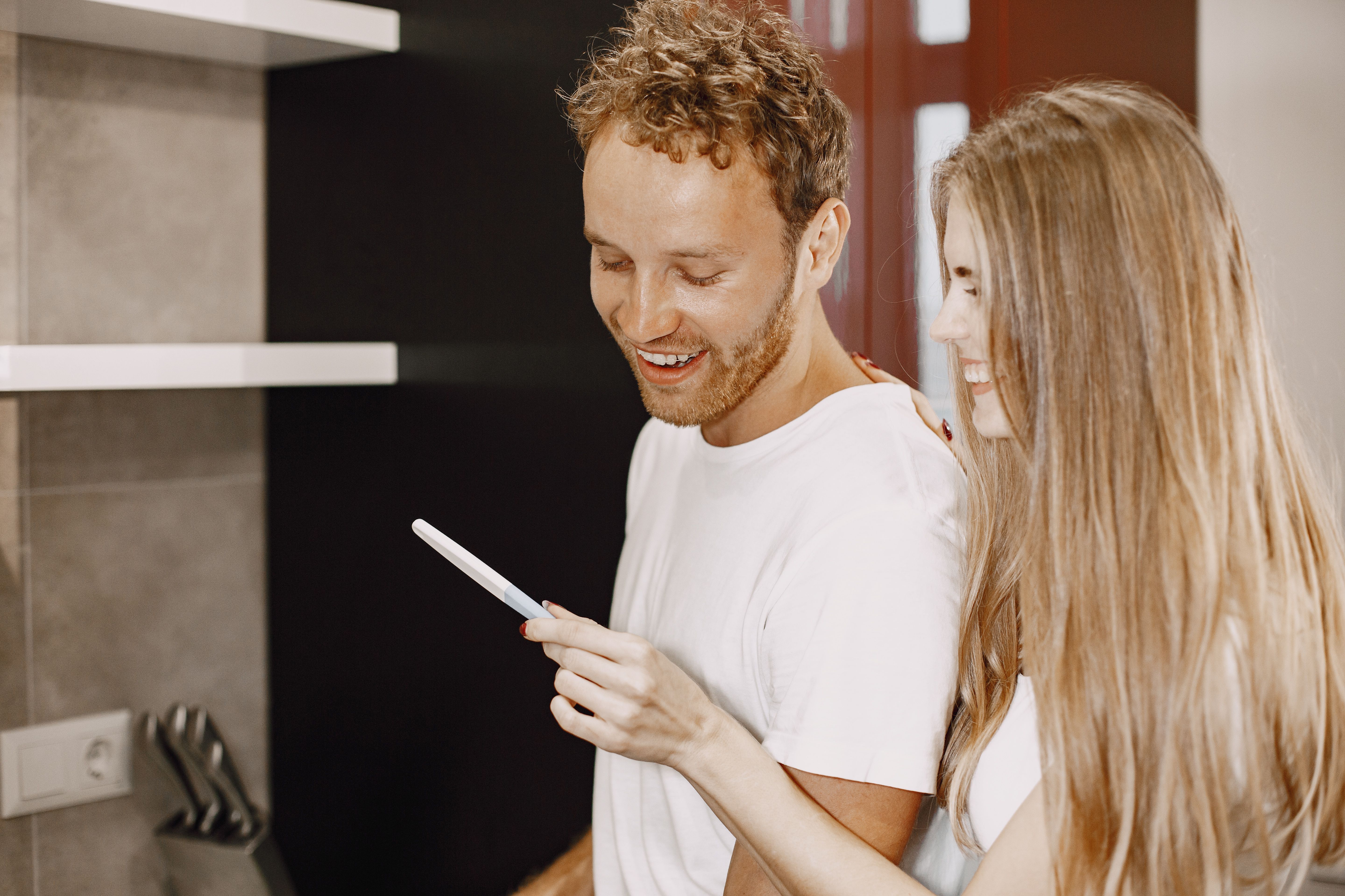 A man smiles next to his wife, who is holding a phone. This image relates to a guy seeing something suspicious on his wife's phone.