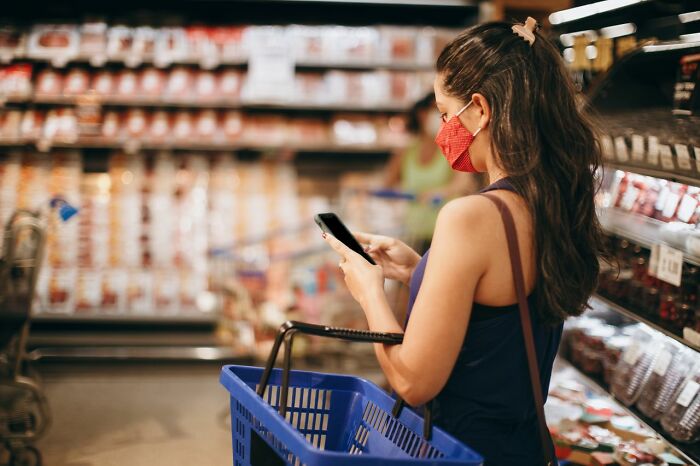 A woman wearing a red mask looks at her phone in a supermarket, feeling her instincts to get away from creeps.