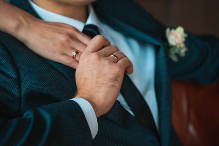 A woman's hand resting on a man's arm, both wearing rings, depicting personal secrets or hidden relationships.