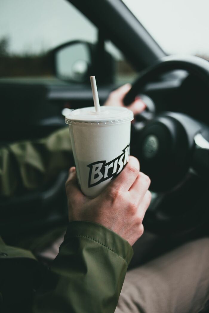 A person holding a disposable cup with the word 'Brisket' written on it while driving, revealing a fascinating everyday moment.