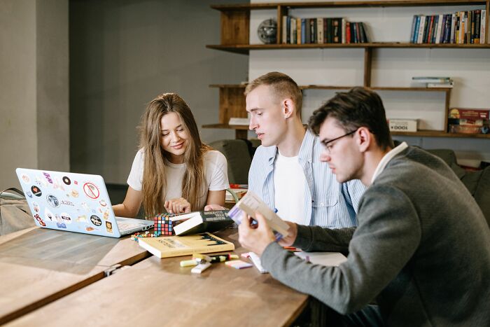 Three young adults engaging in a general knowledge quiz, with a laptop, books, and a Rubiks cube on a table. Three young adults engaging in a general knowledge quiz, with a laptop, books, and a Rubiks cube on a table.