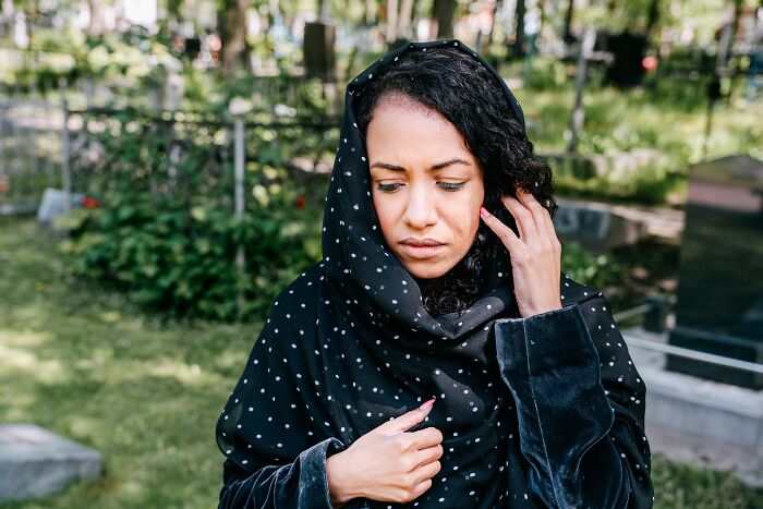 A solemn woman in a dark head covering standing in a graveyard, representing life's unexpected twists and turns.