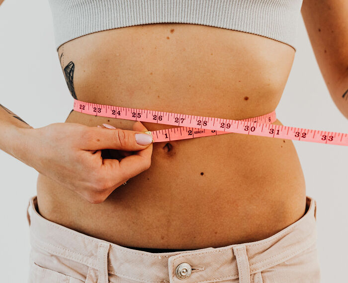 A woman measuring her waist with a pink tape measure. This image relates to clothing sizes and dress experiments. A woman measuring her waist with a pink tape measure. This image relates to clothing sizes and dress experiments.