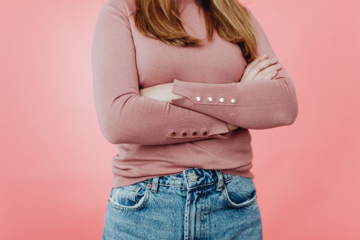 A woman with long hair and a pink sweater, standing with her arms crossed, considering a nanny for a family vacation.