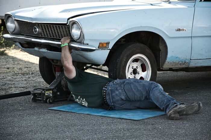 A man in a green shirt and jeans lies on a blue mat, working under the front end of a light blue vintage car, illustrating an unusual way people lost lives.