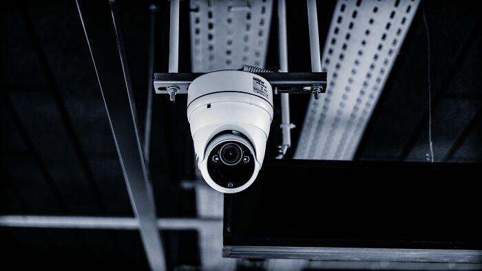 A white surveillance camera hangs from the ceiling, capturing office stories.