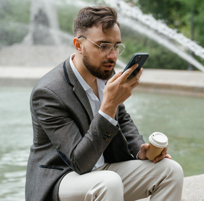 Man on a modern dating app, holding phone and coffee by a fountain, after an unhinged voice memo. Man on a modern dating app, holding phone and coffee by a fountain, after an unhinged voice memo.