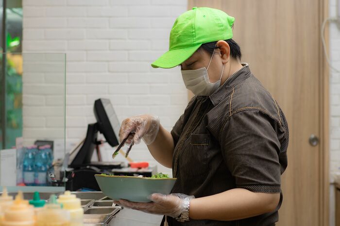 A restaurant worker, wearing a mask and gloves, preparing food, reflecting on dumb reasons people got fired.