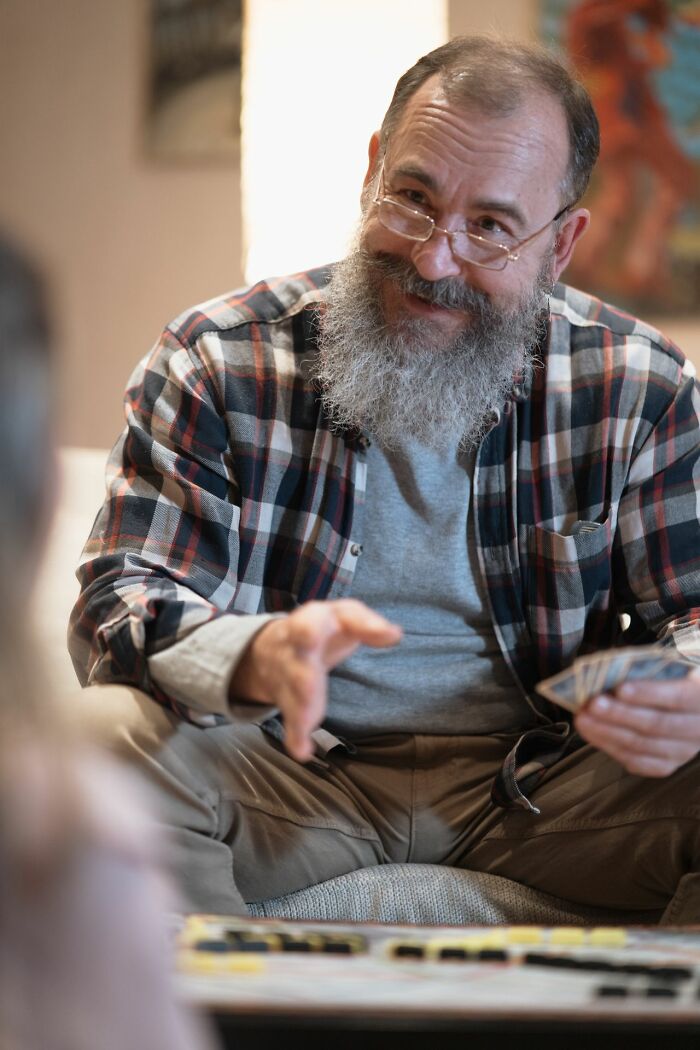 A smiling man with a beard and glasses plays cards, looking at the camera. This image relates to secrets and how they could do damage.
