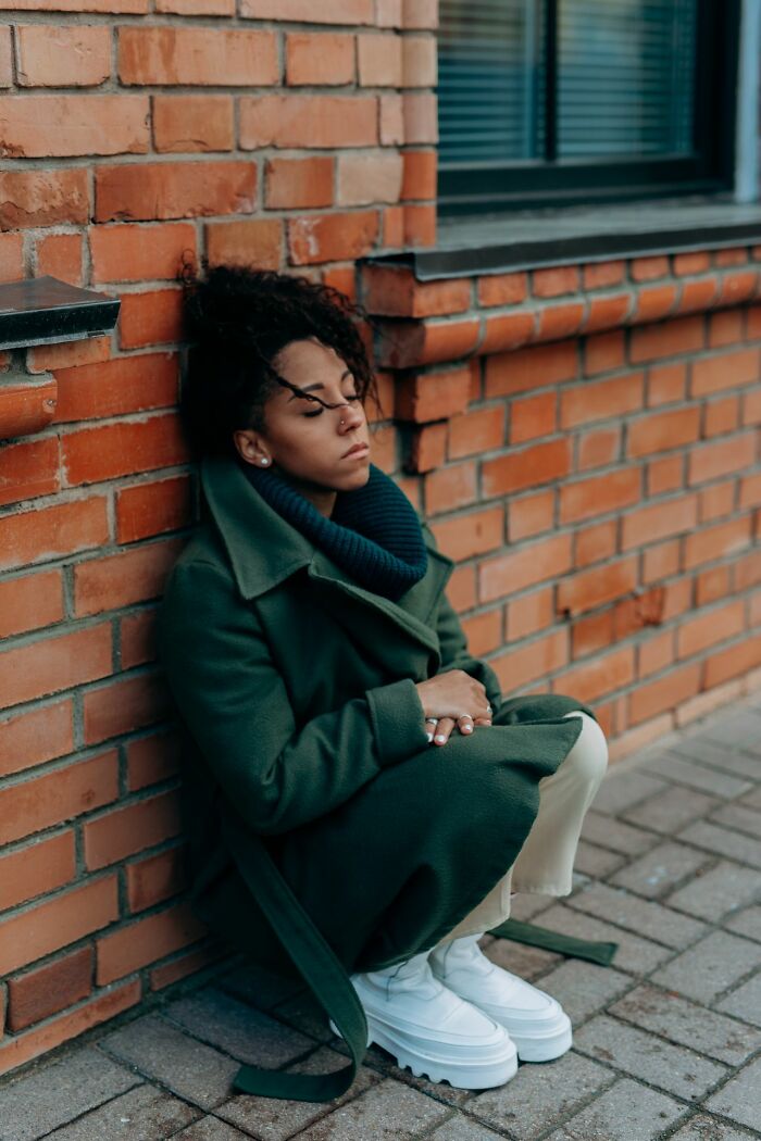 A woman sits against a brick wall, eyes closed, feeling her instincts telling her to get away from creeps.