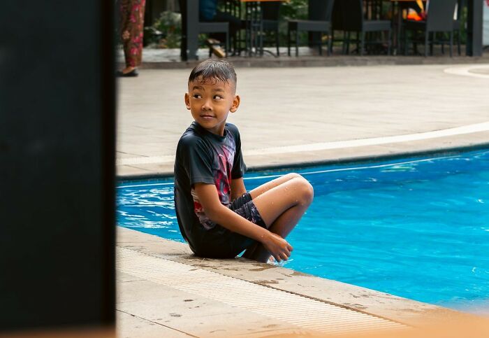 A young boy in swimming trunks sitting by a pool, his expression capturing a moment from a public horror story.