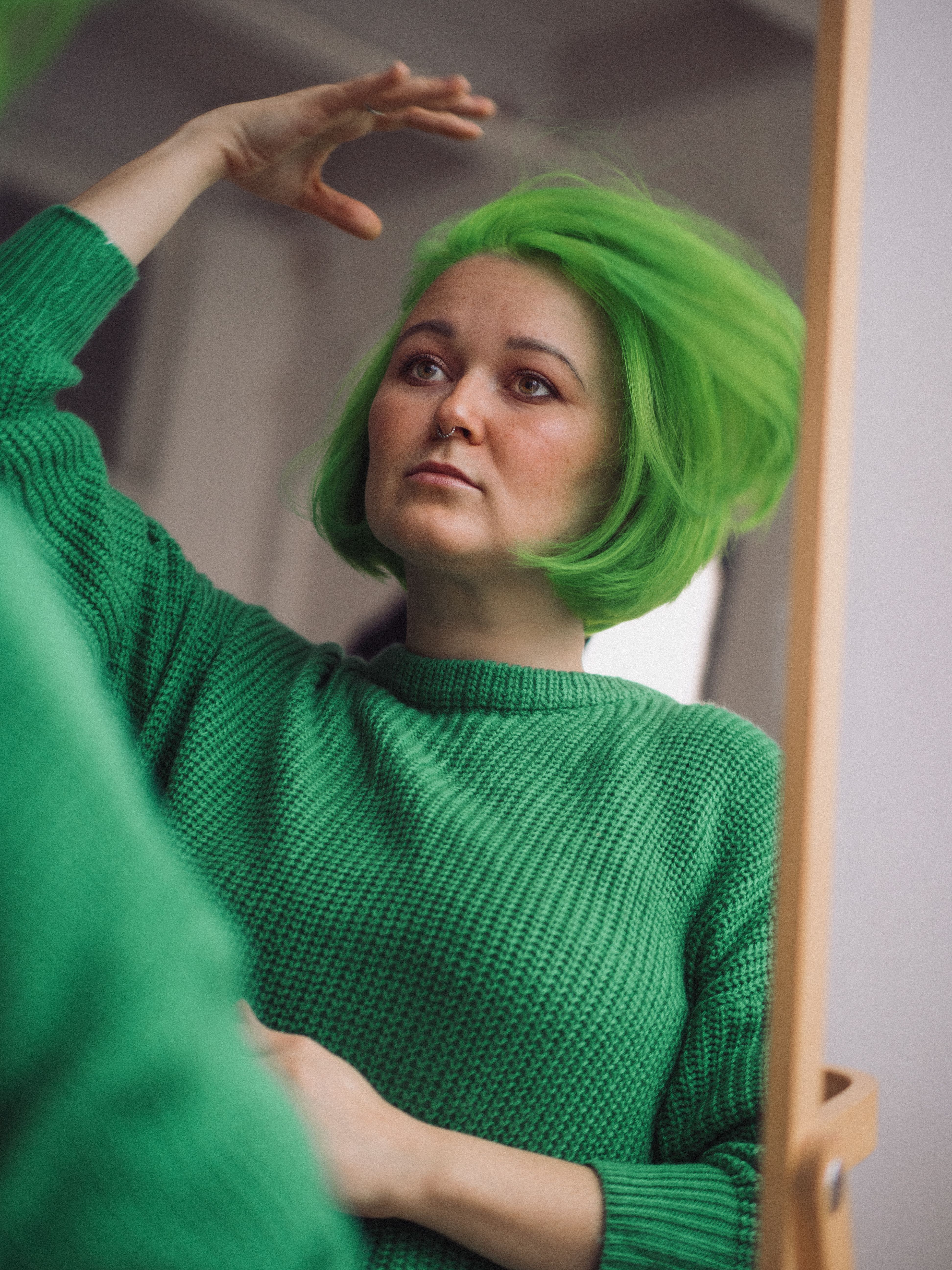 A woman with bright green hair and a matching sweater looks in a mirror, reflecting a scary fixation.