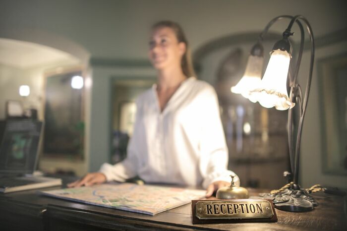 A smiling woman standing behind a hotel reception desk with a bell and a map, likely hearing secrets.