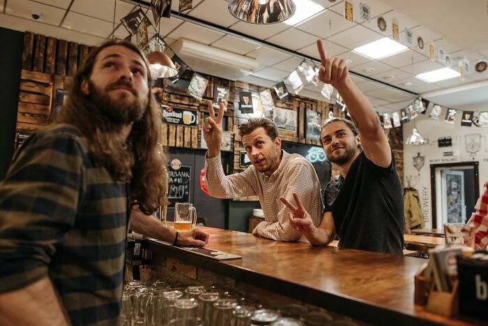 Three men at a bar participating in a pub quiz, testing their general knowledge, with one man raising his hand to answer. Three men at a bar participating in a pub quiz, testing their general knowledge, with one man raising his hand to answer.