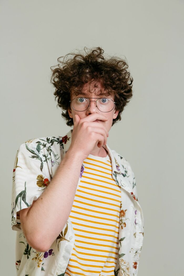 A man with curly hair and glasses, looking surprised with his hand over his mouth, having heard secrets.