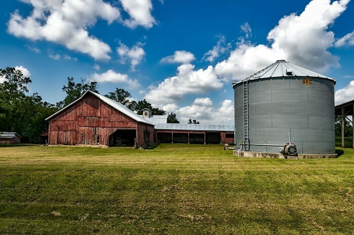 An old red barn next to a large metal grain silo under a bright blue sky with white clouds, highlighting unusual ways people lost lives.