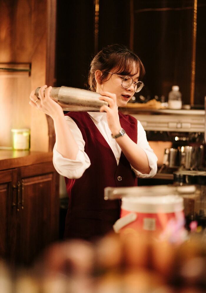 A bartender in a maroon vest shaking a cocktail shaker, showing skill among entitled friends.