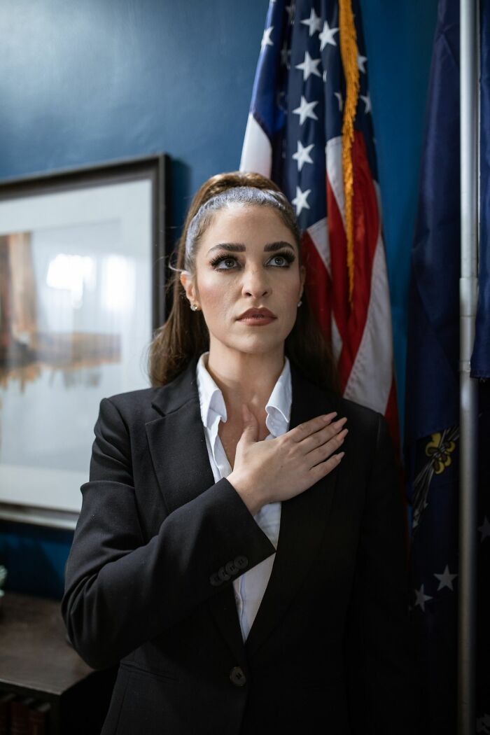 A woman with her hand over her heart in front of an American flag, displaying American habits.