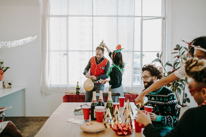 A group of people at a party, one man wearing a Christmas vest and blowing up a balloon. They are sharing secrets.