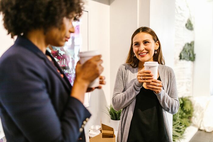 Two women smiling and chatting while holding coffee cups, sharing a casual office story.