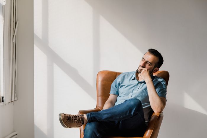A woman creates a plan to make DIL have kids; a man in a blue shirt sits in a brown chair, pondering, in dappled sunlight.