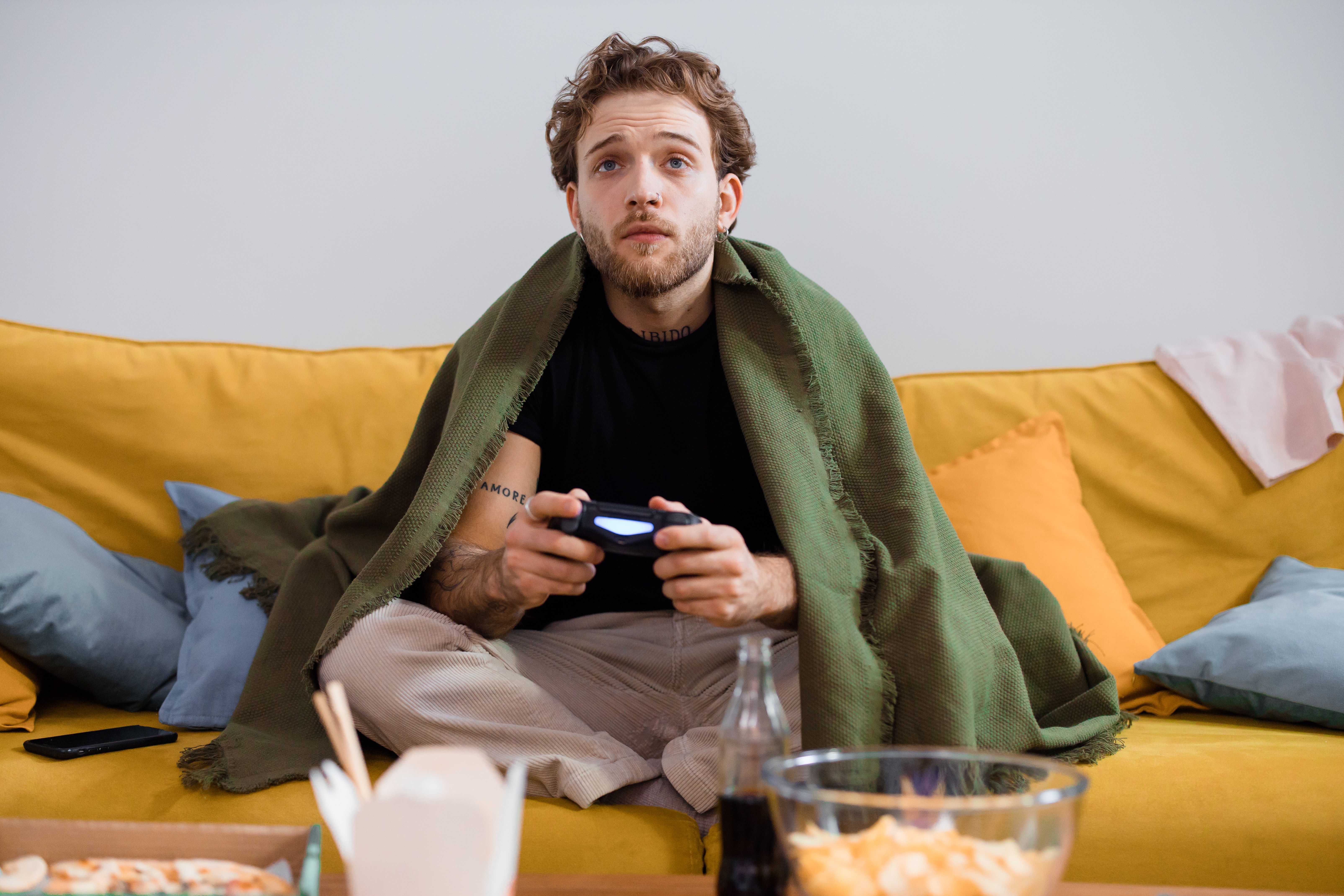A person with a beard, wrapped in a green blanket, sits on a yellow couch holding a video game controller, sharing a simple trick that changed their life.