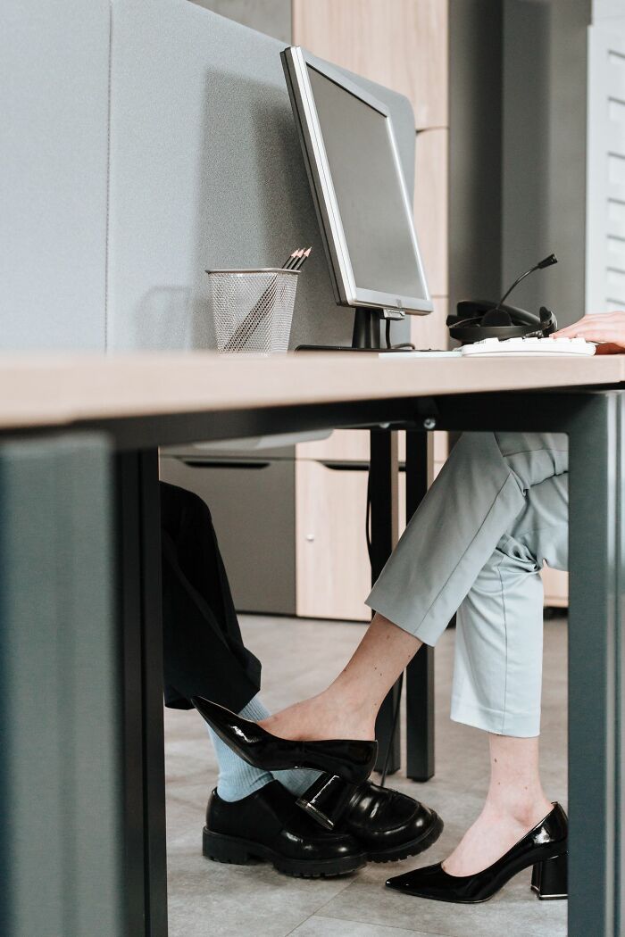 A pair of feet, one in a black shoe and one bare, touching under an office desk, illustrating people's secrets.