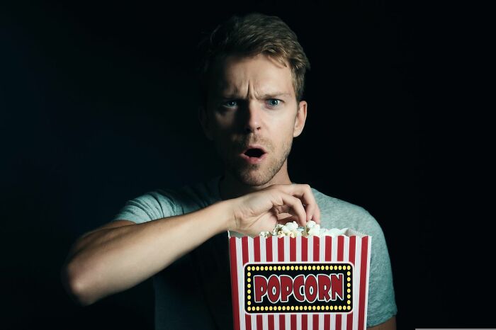 A man with a surprised expression eating popcorn, reacting to shocking secrets he wasn't supposed to know.