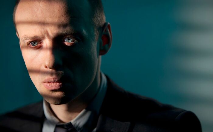A man with striking blue eyes, shadowed by blinds, creating an unsettling look of deeply evil creeps.