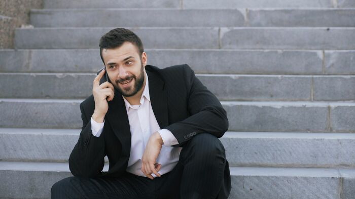 A man in a suit sitting on stairs, smiling while talking on a phone, perhaps discussing secrets and their damage.