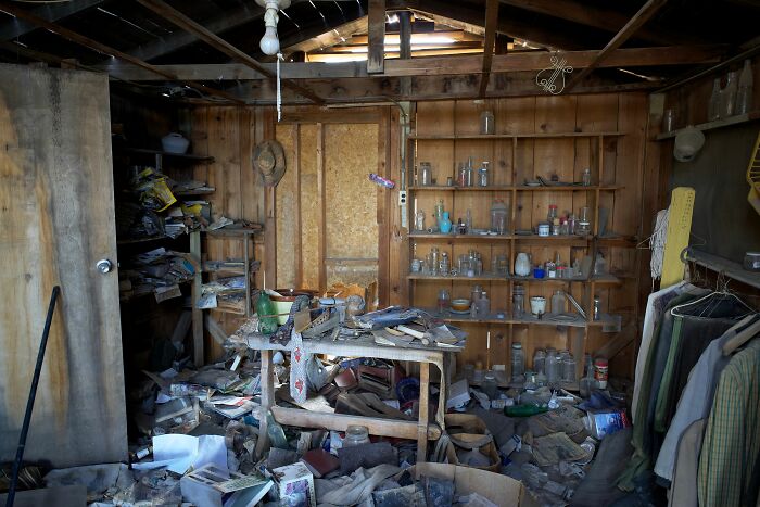 A messy room, possibly a shed or workshop, filled with various items and shelves of bottles, showing a friend's house.