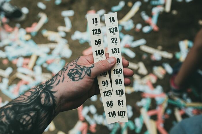 A tattooed hand holding two strips of numbered raffle tickets against a blurred background of confetti, a fascinating detail.