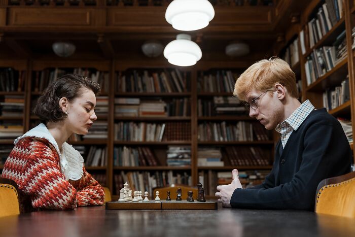 Two people playing chess, testing their general knowledge and high IQ in a library setting, with bookshelves in the background.