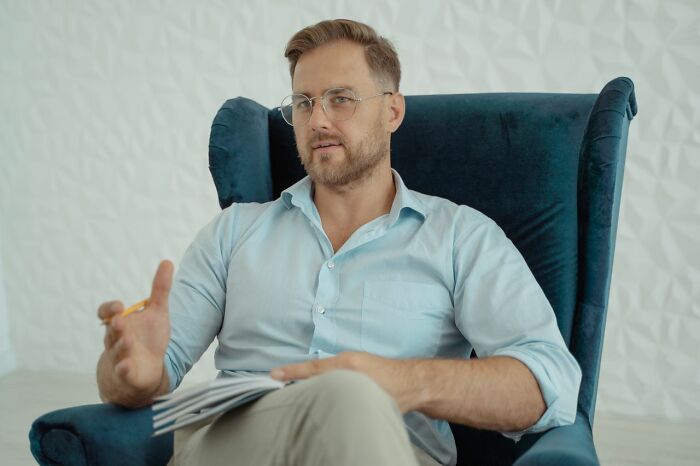 A male therapist in glasses and a light blue shirt, gesturing while holding a notepad, illustrating unhinged therapist moments.
