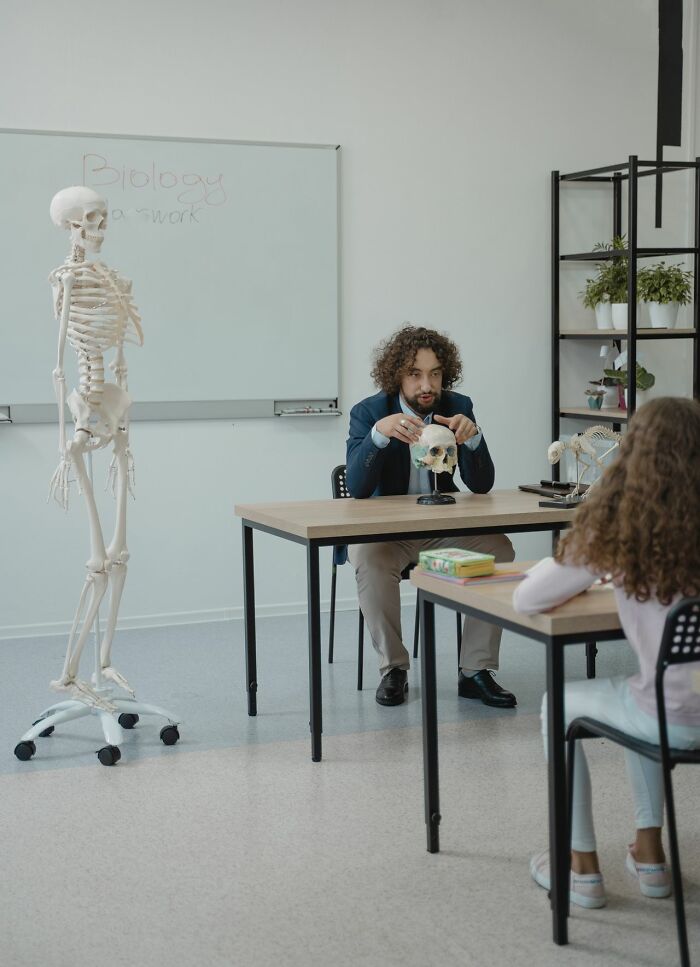 Biology class with a teacher holding a skull, a student, and a skeleton, symbolizing deeply evil education.