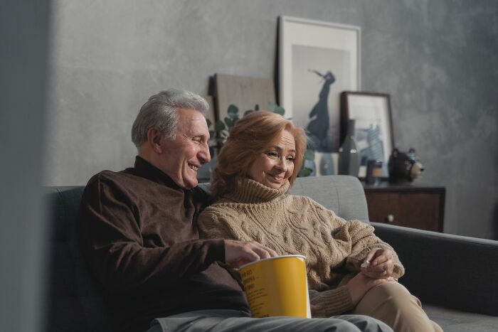 An older couple smiles on a couch with popcorn, showing a peaceful family setting and different versions of normal.